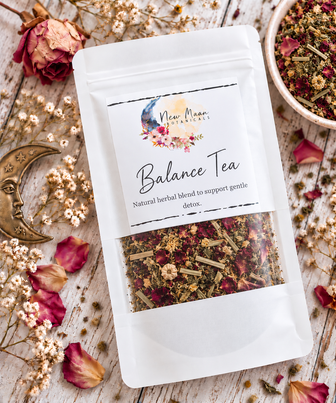 A white stand-up pouch of New Moon Herbs 'Balance Tea' is photographed from above on a rustic whitewashed wooden surface, surrounded by dried roses, petals, a small bowl of loose tea, and a brass moon ornament. The tea blend is visible through a clear window near the bottom of the pouch.