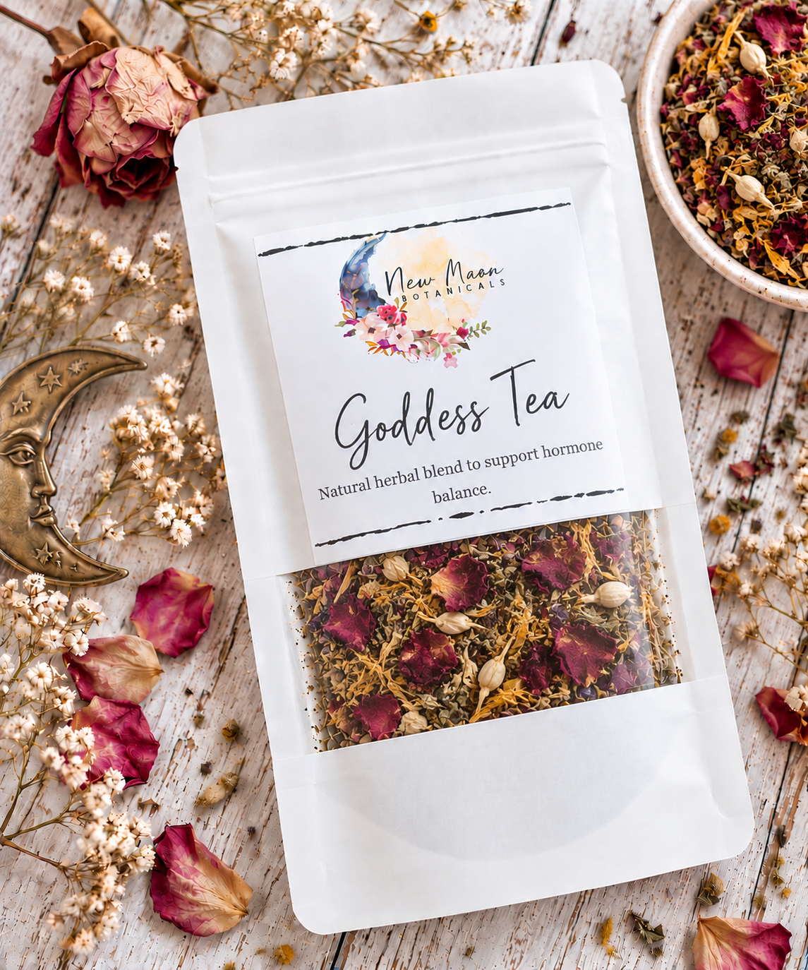 A white stand-up pouch of New Moon Herbs & Teas Goddess Tea is displayed on a rustic white wooden surface surrounded by dried roses, baby's breath, a brass moon ornament, and a small bowl of loose herbal tea blend.