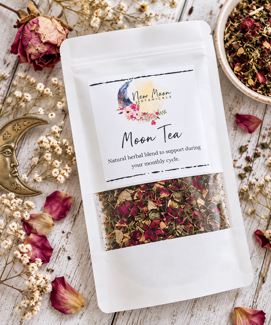 A white stand-up pouch of Moon Tea herbal blend by New Moon Apothecary, displayed on a rustic white wood surface with dried roses, small white flowers, a brass crescent moon ornament, and a bowl of loose tea.