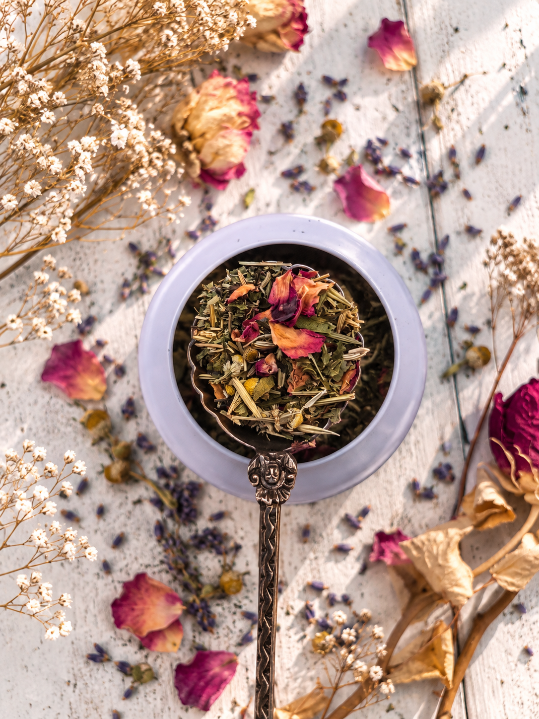 A top-down styled shot of a light blue ceramic bowl filled with loose herbal tea blend containing dried rose petals, surrounded by scattered dried flowers and petals on a white textured surface.