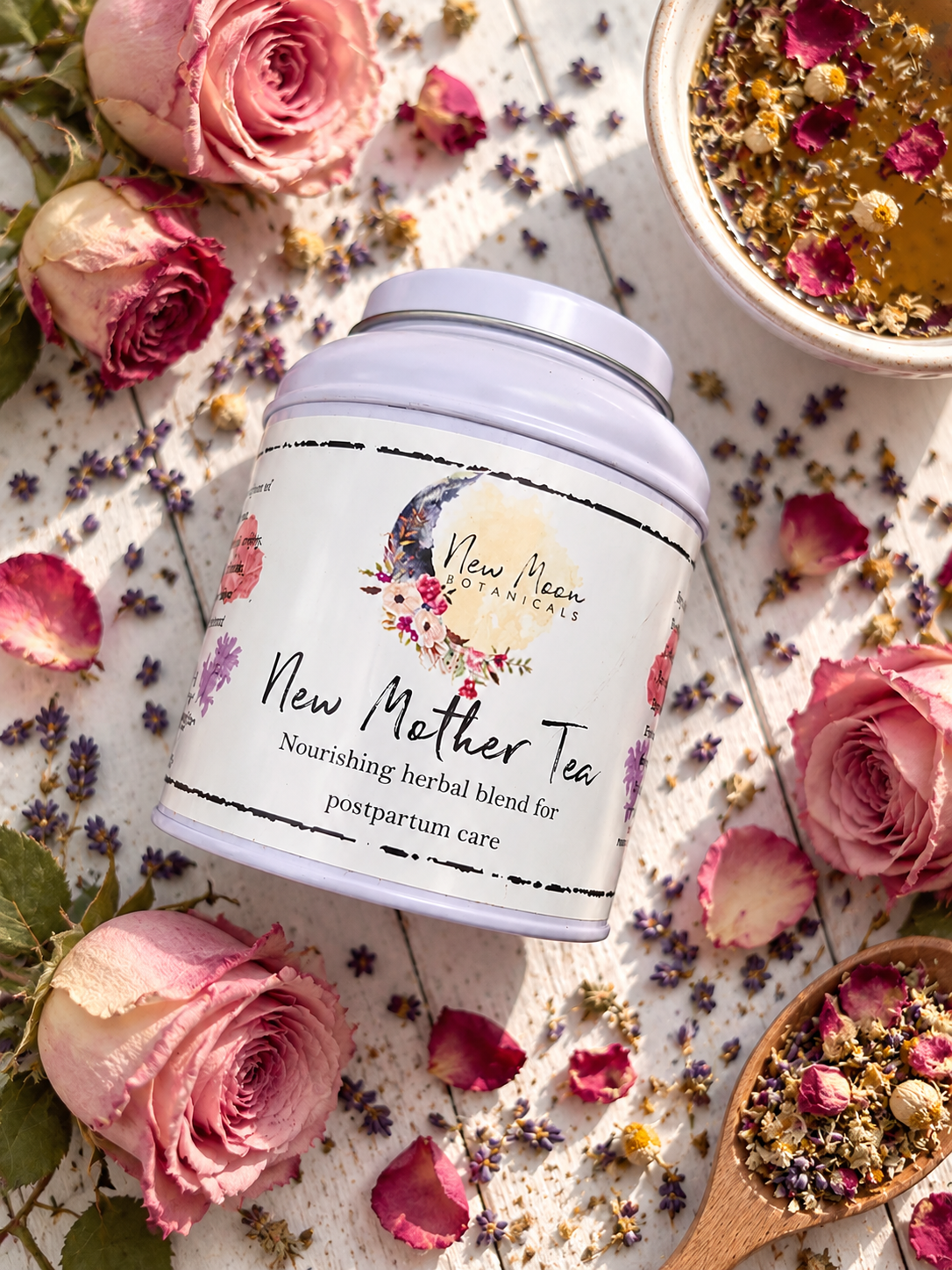 A white cylindrical tin of New Moon Botanicals New Mother Tea is styled in a flat lay with pink roses, dried lavender, and a wooden spoon of herbal blend on a white textured surface.