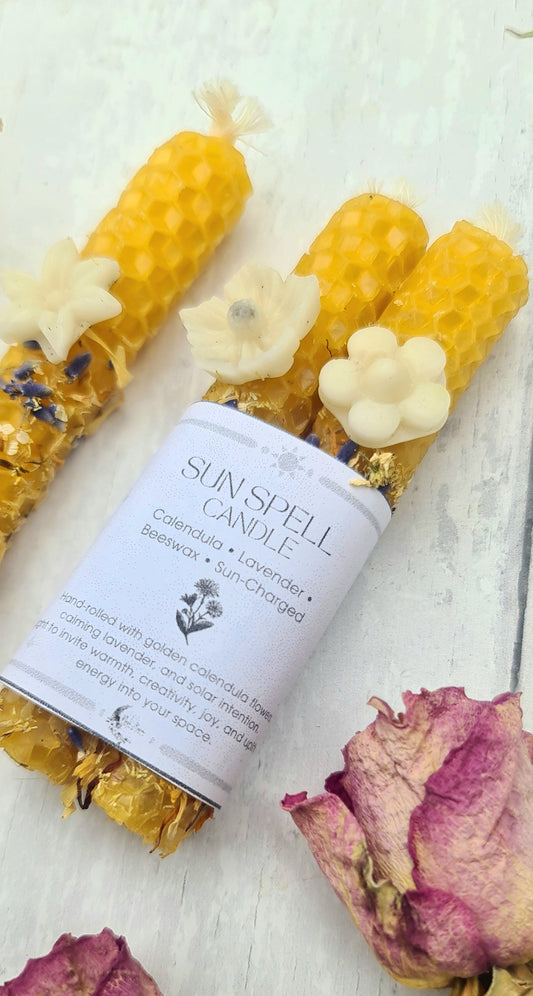 A handmade beeswax candle wrapped in a white paper label reading 'SUN Spell CANDLE' and 'Lavender & Beeswax', styled with dried roses and yellow honeycomb wax beads on a white wooden background.