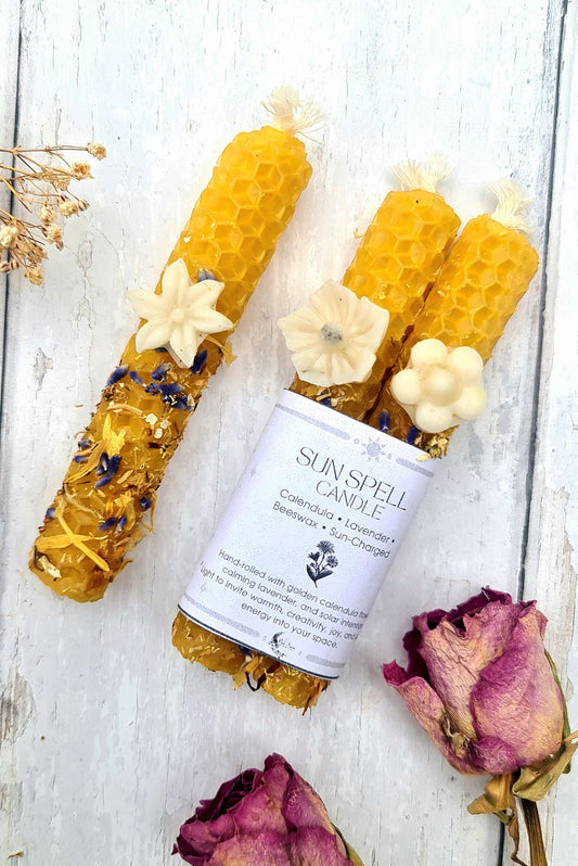 Three yellow honey beeswax candles with dried flowers and herbs are arranged on a white wooden background next to a white paper label. Two pink dried roses are placed near the bottom right.